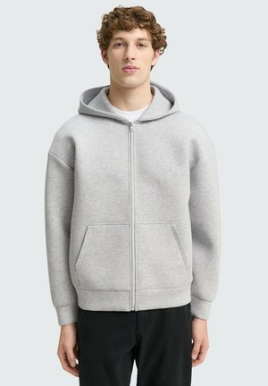 Felpa grigia con zip realizzata in tessuto morbido. Presenta un cappuccio spazioso, due tasche frontali e polsini a costine. Indossata sopra una t-shirt bianca.