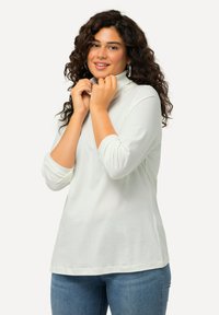 Weißes Langarmoberteil mit hohem Ausschnitt und umgeschlagenen Manschetten, aus einem weichen, glatten Material, kombiniert mit blauen Jeans.