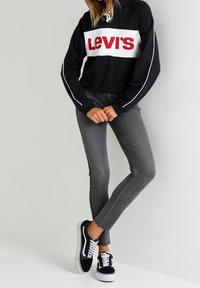 Felpa nera con banda bianca recante il logo rosso "Levi's", abbinata a jeans grigi slim-fit e sneakers basse bianche e nere.