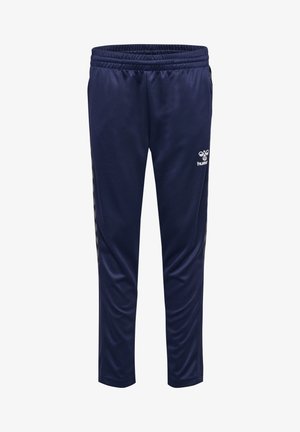 Junge steht mit den Händen in den Taschen, trägt einen dunkelblauen Hummel Trainingsanzug und schwarze Sneakers vor einem einfarbigen hellen Hintergrund.
