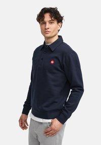 Felpa blu navy con design a colletto, caratterizzata da un logo rosso sul petto, polsini a costine e un look a strati sopra una camicia bianca.