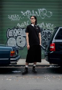 Tricou polo negru cu guler alb, pantaloni scurți negri până la genunchi, cizme negre și șosete albe, stând între două mașini parcate. Fundal cu graffiti verde.