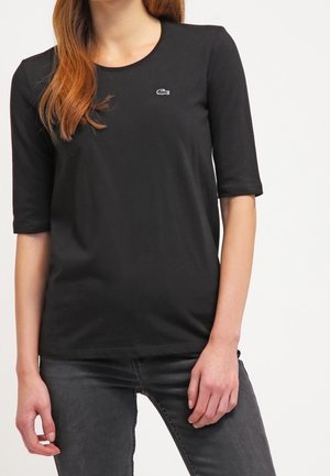 Woman wearing a black short-sleeve top with a small crocodile logo on the chest and dark gray jeans, standing with one hand relaxed.