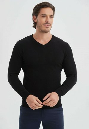 Pull en tricot noir avec un col en V, des manches longues et une coupe ajustée. Porté sur un pantalon bleu foncé, montrant une texture lisse.