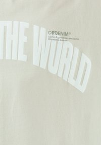 Light beige fabric t-shirt featuring bold white text "THE WORLD" and smaller green logo "DRDENIM," with a smooth texture.