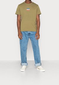 Khaki puuvillapaita, jossa on pieni punainen ja sininen Levi's-logo, yhdistettynä vaaleansinisiin farkkuihin ja kukkakoristeisiiin valkoisiin kenkiin.