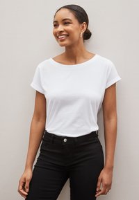 Femme aux cheveux foncés en chignon souriante, portant une chemise blanche à manches courtes et un pantalon noir, debout devant un fond clair uni.