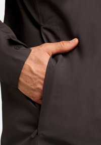 Veste pour homme gris foncé en tissu lisse et texturé ; présente une poche latérale avec une main partiellement visible et un détail de poignet en velcro.