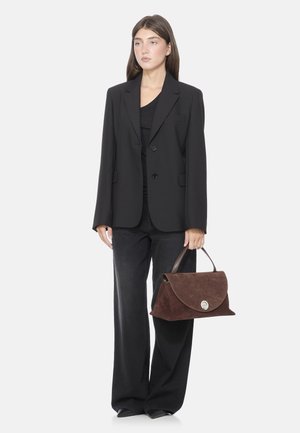 Borsa in suede marrone con design a patta, chiusura circolare in argento e manico in pelle. Indossata con un blazer nero e jeans a gamba larga.