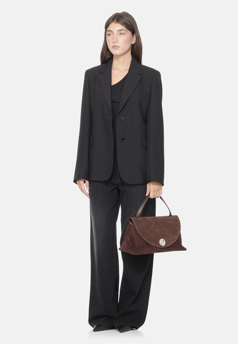 Borsa in suede marrone con design a patta, chiusura circolare in argento e manico in pelle. Indossata con un blazer nero e jeans a gamba larga.
