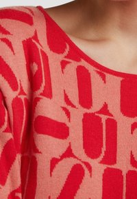 Sweter dzianinowy z szerokim dekoltem, zdobiony abstrakcyjnym wzorem w odcieniach czerwieni i jasnego różu. Miękka tekstura i luźny krój podkreślają design.