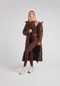 Abrigo acolchado marrón con un corte largo y relajado; superpuesto sobre un top corto de chocolate y leggings a juego; combinado con zapatillas blancas.