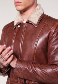Uomo con giacca di pelle marrone e colletto in pelliccia beige chiaro, mani con piccoli tatuaggi vicino al petto, senza volto visibile.