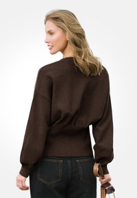 Brązowy dzianinowy sweter o luźnym kroju, z balonowymi rękawami, ściągaczem na dole i gładką teksturą, w połączeniu z ciemnymi dżinsami i okularami przeciwsłonecznymi.