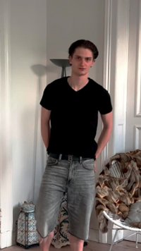 Camiseta preta de mangas curtas, calções de ganga cinza desgastados, de pé numa sala com paredes brancas, base de lâmpada com padrão e cadeira trançada.