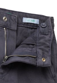 Pantalones gris oscuro con una cremallera abierta, botón y cierre a presión que muestran una etiqueta azul clara con el texto verde "LILY-JORT."