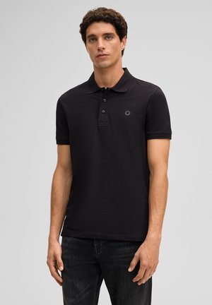 Jonge man staat frontaal, draagt een effen zwarte polo met korte mouwen en donkere spijkerbroek, tegen een neutrale achtergrond.