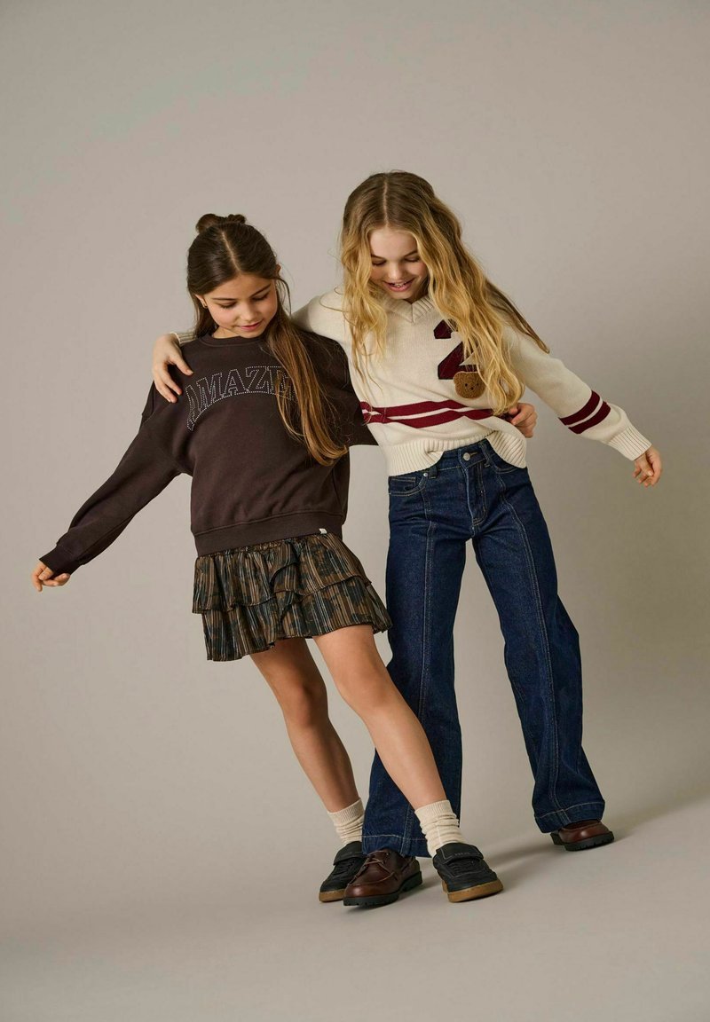 Djevojke nose tamni duksericu s natpisom "AMAZING" i bijeli pulover s bordo prugama. Jedna nosi suknju s volanima, dok druga nosi traperice.