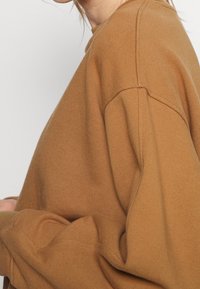 Sudadera marrón con un corte relajado, textura suave y mangas con puños. Presenta un escote redondo y un detalle de costura mínima.