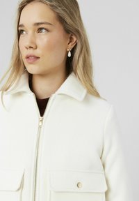 Veste en mélange de laine blanche avec une fermeture éclair sur le devant, un col pointu et deux poches poitrine avec des accents dorés. Portée avec un haut bordeaux en dessous.