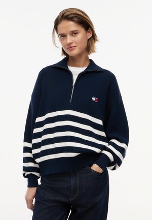 Młoda kobieta z krótkimi włosami, ubrana w granatowo-biały sweter w paski z zamkiem do ćwiartki i ciemne dżinsy, jedna ręka w kieszeni, na białym tle.