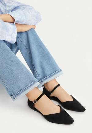 Nogi skrzyżowane, ubrane w jasnoniebieskie dżinsy i czarne, spiczaste baleriny na paski wokół kostki na jednolitym białym tle.