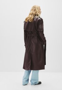 Cappotto trench in finta pelle marrone con cintura in vita, spalline sulle spalle e vestibilità ampia. Abbinato a jeans svasati azzurri chiari.