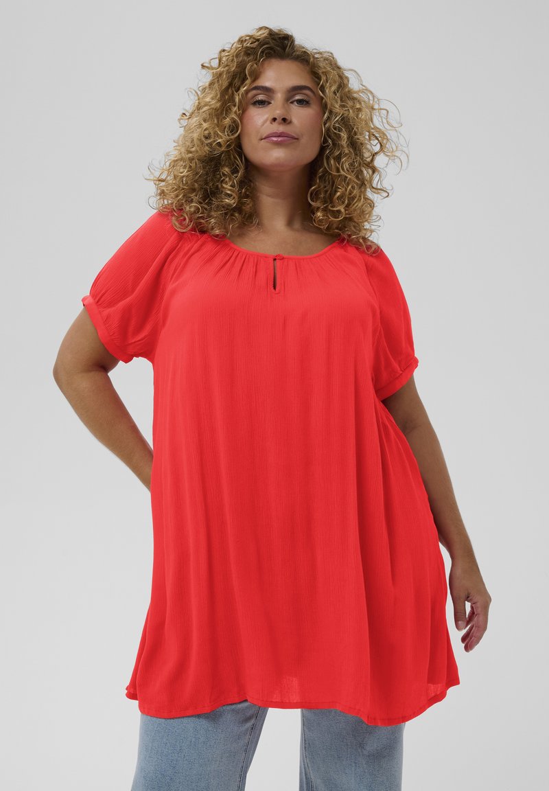 Femme aux cheveux bouclés portant une tunique ample de couleur rouge vif à manches courtes et un jean bleu clair, debout devant un fond uni.