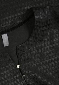 Svart strukturerad blus med liten krage, framknäppning och ett vävt schackrutsmönster för extra detalj. Mjuk tygfinish.