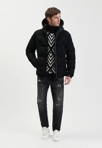 Zwarte puffer jas met een gestructureerd ruitpatroon, gecombineerd met een zwart-wit gestreepte trui, versleten donkere spijkerbroek en witte sneakers.