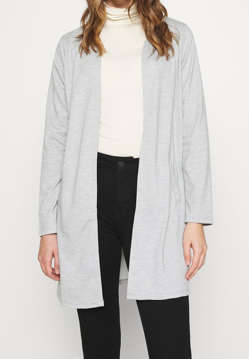 Cardigan long gris clair à coupe décontractée, fabriqué en tissu doux et texturé. Il présente une ouverture sur le devant et des poches latérales, porté sur un haut crème.