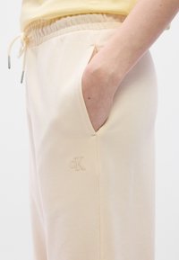 Φωτεινό μπεζ παντελόνι φόρμας από απαλό ύφασμα, με ελαστική μέση, πλαϊνές τσέπες και διακριτικό κεντημένο λογότυπο στο μηρό.