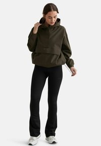 Femme ajustant le cordon d'une veste à capuche oversize vert olive, portant des leggings noirs et des baskets blanches, debout sur un fond blanc.