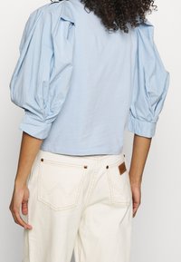 Top de manga abullonada en azul claro con textura fruncida en los hombros; longitud cropped. Combinado con pantalones blancos con detalles en marrón y bolsillos en zigzag.