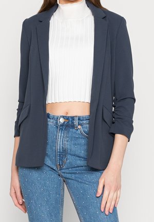 Woman wearing a cropped white ribbed sweater, navy blazer with rolled sleeves, and blue high-waisted jeans decorated with small white dots.