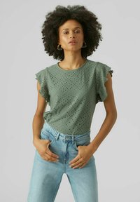 Top in cotone verde con maniche a rouches, caratterizzato da motivi a occhiello e scollatura rotonda, abbinato a jeans azzurri a vita alta.