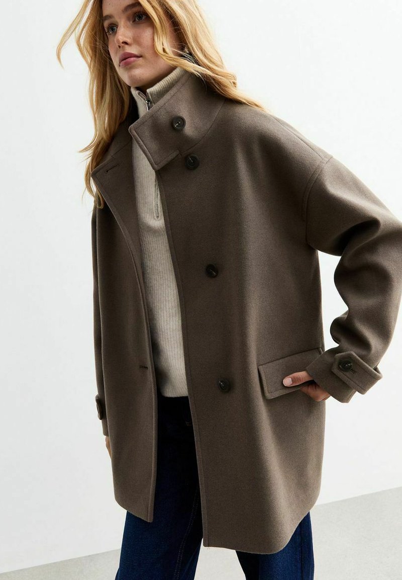Cappotto marrone in misto lana con colletto alto, bottoni doppi, ampie tasche laterali e vestibilità rilassata, indossato sopra un maglione leggero.