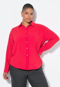 Model with short hair poses confidently in a bright red shirt and dark pants, showcasing a casual yet stylish look against a white background.