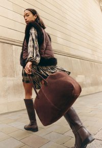 Donna che cammina sul marciapiede con una grande borsa marrone, indossa un vestito a quadri, un gilet scuro con bordo in pelliccia e stivali al ginocchio.