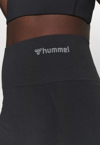 Zbliżenie na talię w czarnych legginsach z wysokim stanem z szarym logo marki "hummel" na pasie.