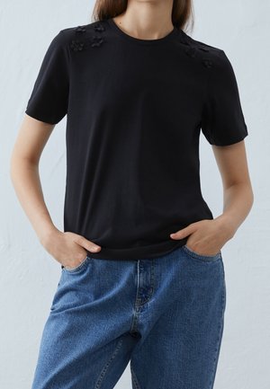 Czarny t-shirt z krótkim rękawem ozdobiony czarnymi aplikacjami w kształcie kwiatów na ramionach; miękka tekstura, klasyczny okrągły dekolt, zestawiony z niebieskimi dżinsami.