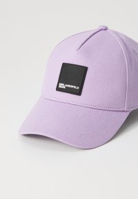Levanduľová baseballová čiapka vyrobená z textúrovaného materiálu, s čiernou štvorcovou nálepkou s nápisom "KARL LAGERFELD JEANS" v bielej farbe na prednej strane.