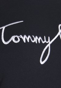 Tela negra con un patrón de punto texturizado que presenta un gran texto bordado en blanco que dice "Tommy" en una fuente cursiva.