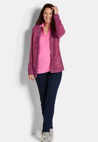 Lächelnde Frau steht mit der Hand im Haar, trägt ein pinkes Shirt, lila Cardigan, dunkle Hose und hellen Sneakern vor einem schlichten Hintergrund.