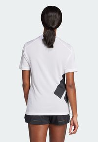 Camiseta atlética blanca con textura de malla, con acentos negros y un diseño distintivo. Combinada con pantalones cortos oscuros. Vista desde atrás.