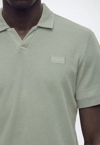 Κοντινό πλάνο ενός ατόμου που φοράει ανοιχτοπράσινο πόλο με υφή, γιακά και ένα διακριτικό ορθογώνιο λογότυπο στο στήθος.