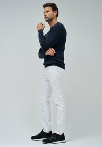 Sweater a coste blu navy, pantaloni slim-fit bianchi e sneakers nere. Modello in piedi con le braccia incrociate, catturato di profilo su uno sfondo neutro.