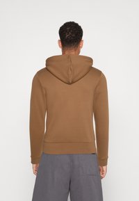 Bruine hoodie met een trekkoordcapuchon, aansluitende manchetten en zoom. Zacht stof, eenvoudig ontwerp en een relaxte pasvorm. Draag het over grijze shorts.