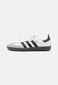 White leather sneakers with black stripes, grey suede toe cap, grey rubber sole, and blue accent on the tongue. Gold "Samba" detailing.