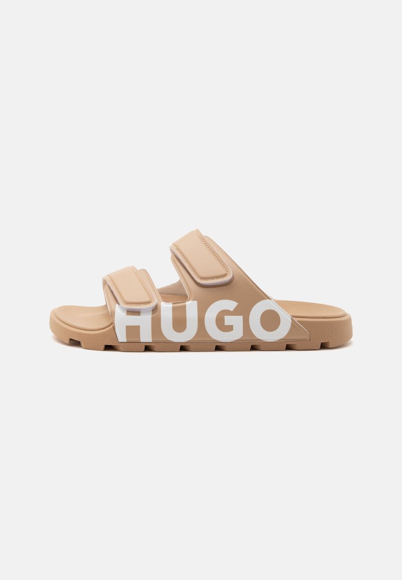 Sandale beige à enfiler avec deux brides réglables et une semelle épaisse, avec un grand texte blanc "HUGO" sur le côté.